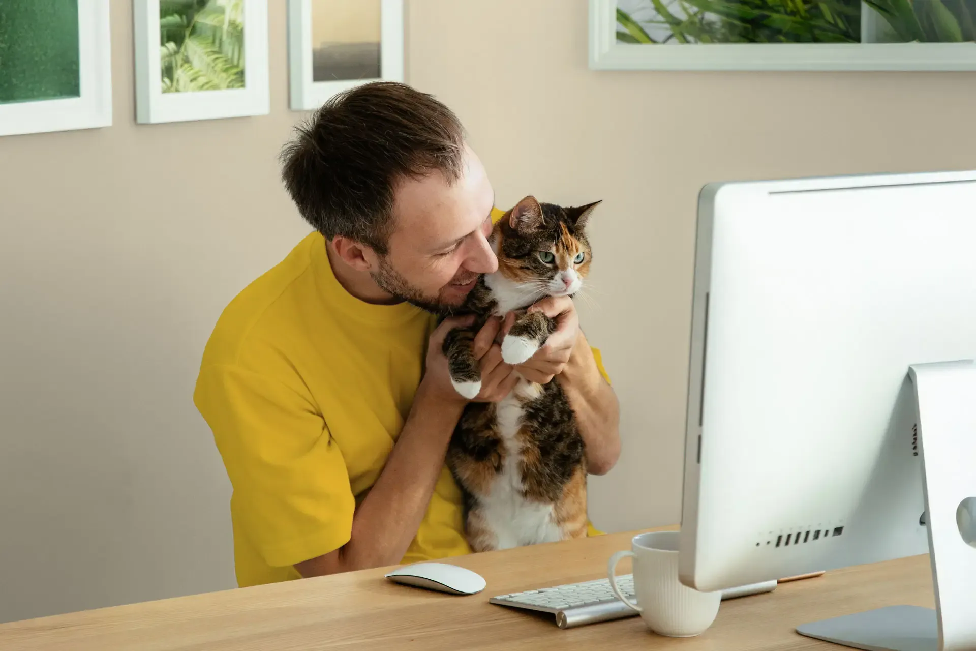 A man in a bright yellow shirt sits at his desk, lovingly holding his cat
