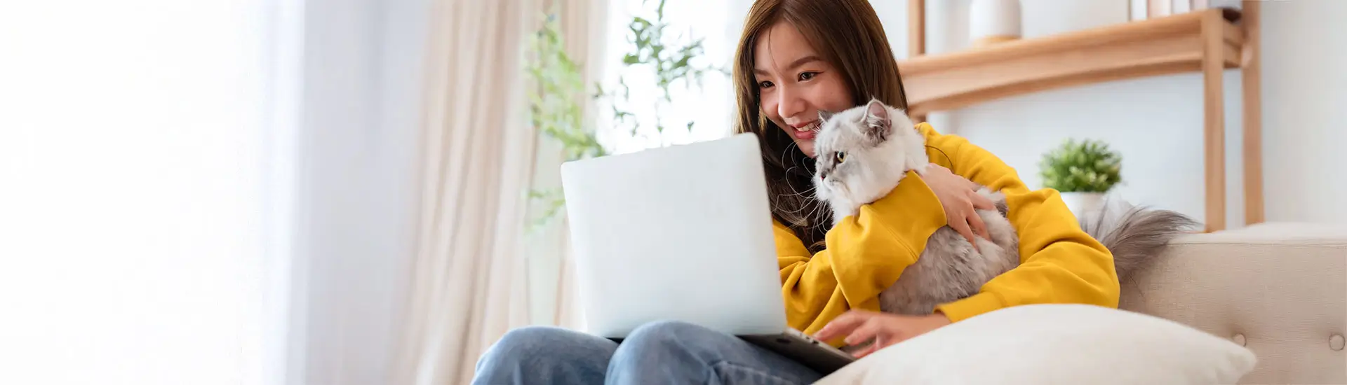 A cheerful young woman sits in a comfortable chair, hugging her cat. She smiles while working at her laptop.