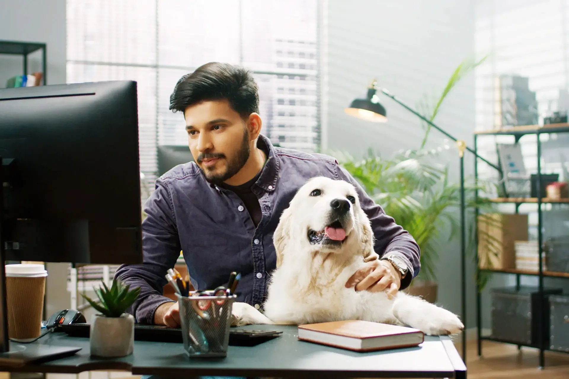 Bearded man sits at his desk and works while his happy dog sits close to him