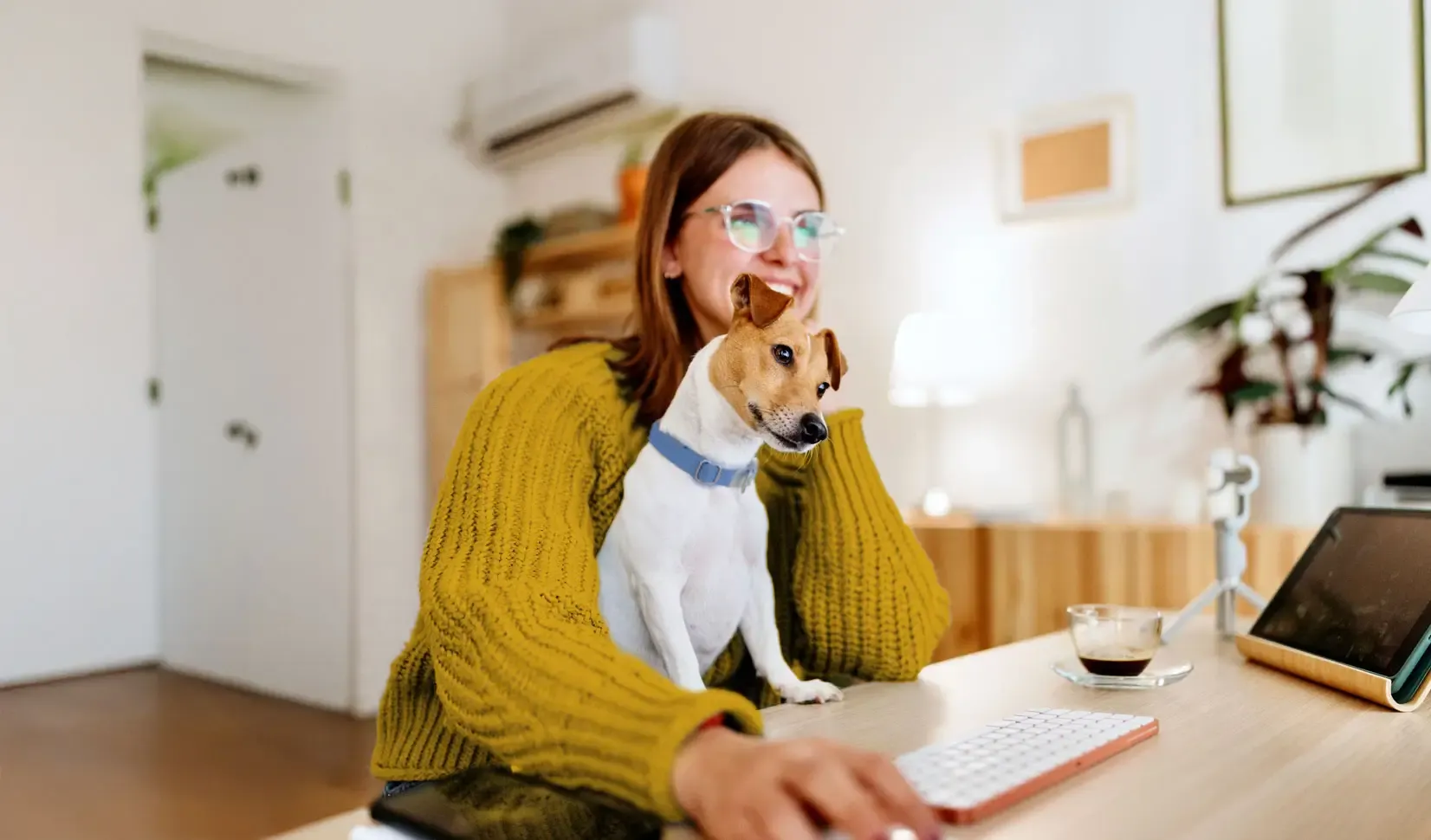 A smiling woman sits at her desk computer with a cute dog in her lap